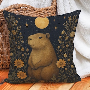 May include: Square decorative pillow featuring a capybara illustration. The design includes a brown capybara, yellow flowers, a full moon, and stars against a dark blue background. The pillow is placed on a wooden surface near a wicker basket and a white blanket.