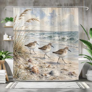 May include: A shower curtain featuring a watercolour painting of three sandpipers walking along a sandy beach. The scene includes tall beach grass on the left, scattered seashells on the sand, and ocean waves rolling towards the shore under a cloudy sky.