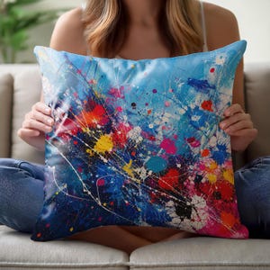 Op de afbeelding: Een vierkant sierkussen met een levendig abstract ontwerp met een blauwe achtergrond bedekt met veelkleurige verfspatten, waaronder helderrode, gele, roze en witte druppels en strepen die een dynamisch en energiek visueel patroon creëren.