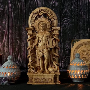 Puede incluir: Una estatua de madera de un hombre con un halo de rayos de sol, sosteniendo una lira, flanqueado por serpientes. Dos candelabros decorativos y una vela encendida están en primer plano. Cristales de amatista dispersos.