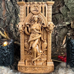 May include: A detailed wooden carving of a woman with a snake wrapped around her, set within an arched frame. The carving features intricate details, including animal heads and owls. The background includes a tree trunk and two black candle holders.