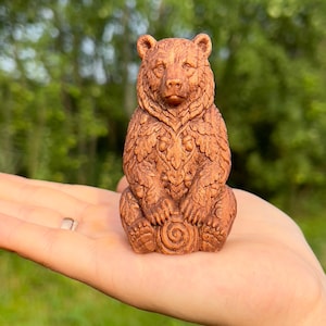May include: A small, brown, hand-carved wooden figurine of a bear sitting in an upright position. The bear's fur is textured with intricate, carved leaf and acorn patterns across its chest and body, with a spiral design visible on its base.