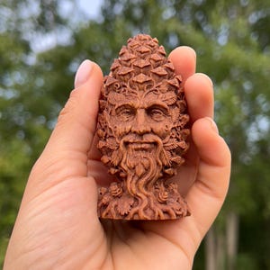 May include: A brown wooden carving shaped like a pinecone with a detailed face of a bearded figure integrated into the texture. The sculpture features intricate leaf and vine patterns surrounding the eyes, nose, and mouth, with a pointed top.