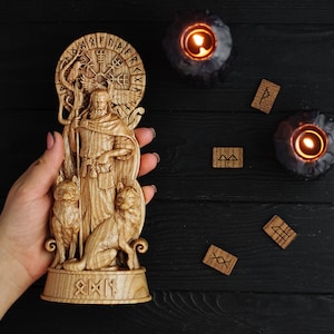 K&ouml;nnte beinhalten: Eine Holzschnitzerei eines nordischen Gottes mit zwei W&ouml;lfen an seiner Seite. Die Schnitzerei ist von einem Kreis aus Runen umgeben und wird in einer Hand vor einem schwarzen Holzgrund gehalten.