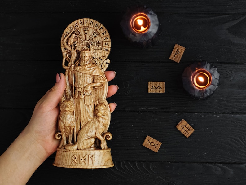 K&ouml;nnte beinhalten: Eine Holzschnitzerei eines nordischen Gottes mit zwei W&ouml;lfen an seiner Seite. Die Schnitzerei ist von einem Kreis aus Runen umgeben und wird in einer Hand vor einem schwarzen Holzgrund gehalten.