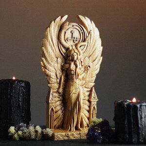 May include: A wooden sculpture of a winged figure with a serpent crown and a pentagram symbol. The figure is standing on a base with two towers. The sculpture is surrounded by black candles and flowers.