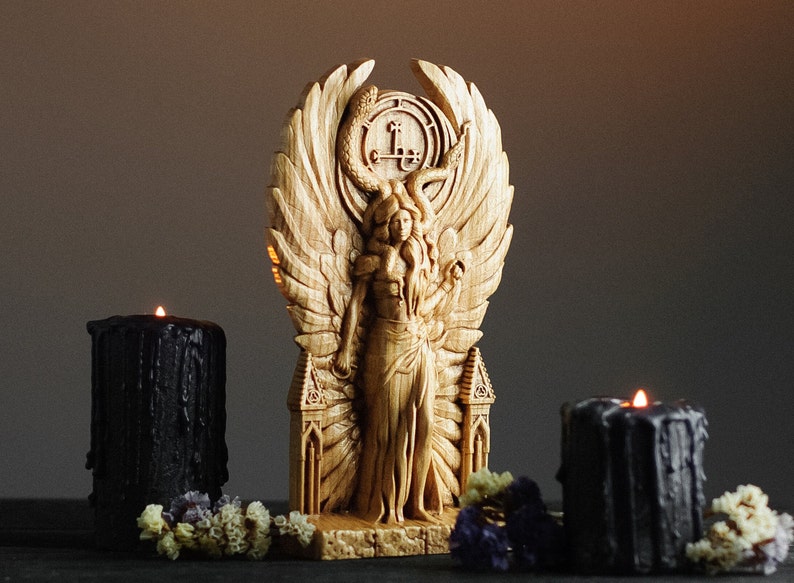 May include: A wooden sculpture of a winged figure with a serpent crown and a pentagram symbol. The figure is standing on a base with two towers. The sculpture is surrounded by black candles and flowers.