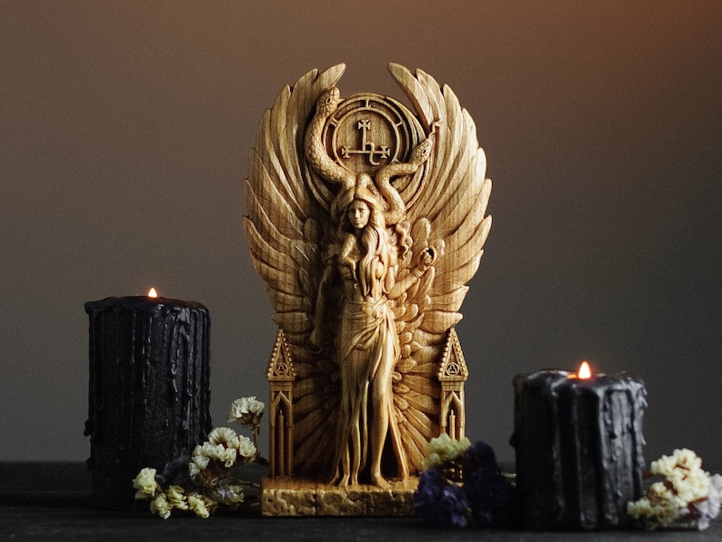 May include: A wooden sculpture of a woman with wings, a snake crown, and a pentagram on her chest. The sculpture is on a wooden base with a carved design. The sculpture is surrounded by black candles and flowers.