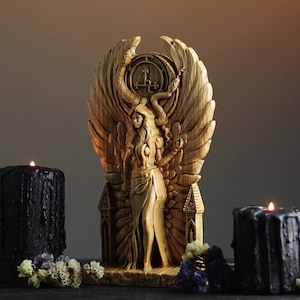 May include: A wooden sculpture of a woman with wings, holding a snake, standing on a throne with a pentagram. The sculpture is surrounded by two black candles.