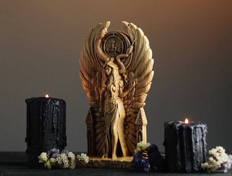 May include: A wooden sculpture of a woman with wings, holding a snake, standing on a throne with a pentagram. The sculpture is surrounded by two black candles.