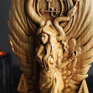 May include: A wooden carving of a woman with wings, a snake, and a symbol. The woman is wearing a crown and a long dress. The carving is painted in shades of brown and gold.