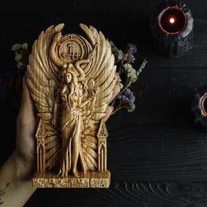 May include: A wooden carving of a winged figure with a snake crown and a pentagram symbol. The figure is standing on a base with a gothic arch design.