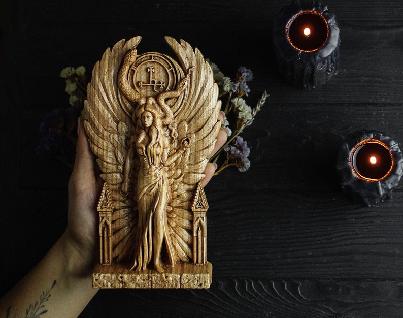May include: A wooden carving of a winged figure with a snake crown and a pentagram symbol. The figure is standing on a base with a gothic arch design.