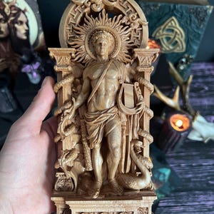 May include: A detailed wooden carving of a classical figure, possibly a deity, with a sunburst halo. The figure holds a lyre and is flanked by columns and serpents. The carving is in a light brown color, with intricate details.