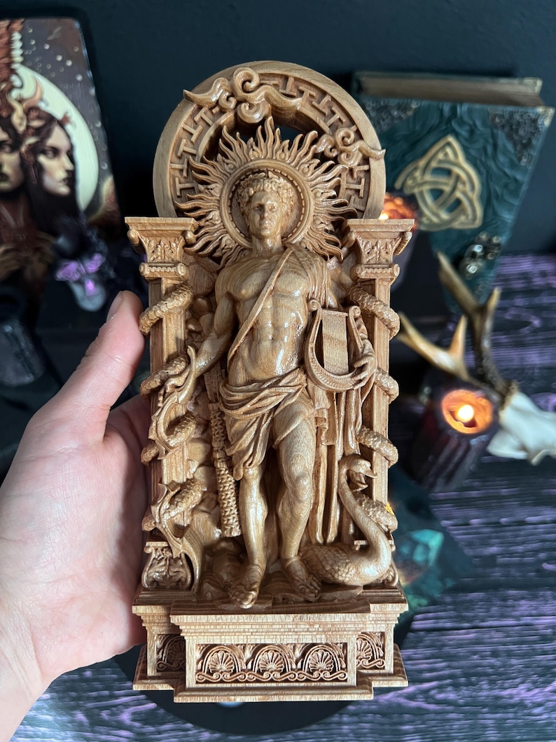 May include: A detailed wooden carving of a classical figure, possibly a deity, with a sunburst halo. The figure holds a lyre and is flanked by columns and serpents. The carving is in a light brown color, with intricate details.