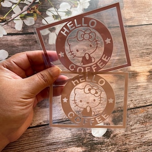May include: Two clear stickers with a Hello Kitty coffee design. The design features the Hello Kitty character inside a brown circle with the words "HELLO" and "COFFEE". The stickers are held against a wooden background.