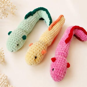 Könnte beinhalten: Drei gehäkelte Axolotl-Stofftiere in Hellgrün, Gelb und Pink. Jedes Stofftier hat einen Körper aus strukturiertem Garn, kleine dunkle Augen sowie kontrastfarbene Kiemen und Rückenflossen entlang des Rückens, die in einer diagonalen Reihe angeordnet sind.