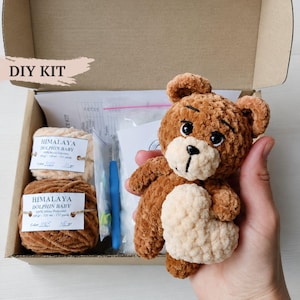 Peut inclure: Un kit DIY dans une boîte en carton contenant deux pelotes de laine Himalaya Dolphin Baby, un crochet bleu, du rembourrage et un patron. Un ours en peluche fini marron et crème tenant un cœur est présenté au premier plan.