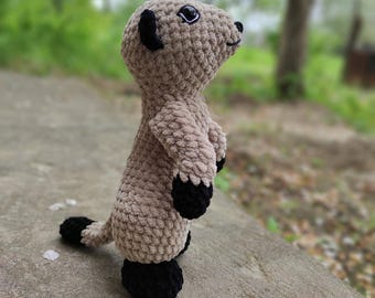 May include: A handmade crochet meerkat plush toy crafted from soft, light tan yarn. The figure features black yarn accents on the ears, paws, feet, and the tip of its tail. It has large, dark, glossy safety eyes and a small, stitched black nose.