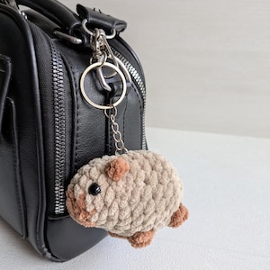 May include: A plush, crocheted capybara keychain in a light tan color with small brown accents on the ears, nose, and feet. The capybara has a round, textured body and a black bead eye, attached to a silver-toned metal keyring and chain.