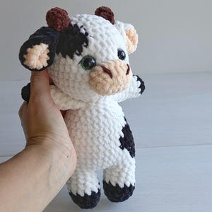 May include: A handmade crochet cow plush toy featuring a white body with black patches, small brown horns, and a light beige muzzle. The stuffed animal has large, glossy black safety eyes and black-tipped hooves on its arms and legs.