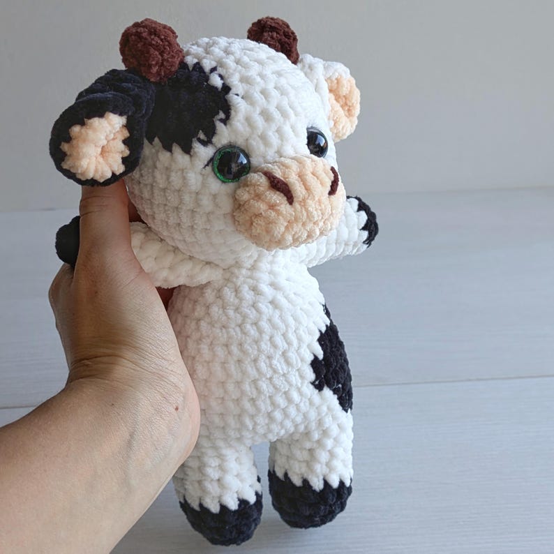 May include: A handmade crochet cow plush toy featuring a white body with black patches, small brown horns, and a light beige muzzle. The stuffed animal has large, glossy black safety eyes and black-tipped hooves on its arms and legs.