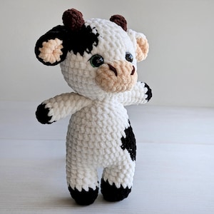 May include: A plush cow toy made of white, black, and brown yarn. The stuffed animal features a white body with black patches, a tan muzzle with dark brown nostrils, small brown horns, and large black eyes. The limbs end in black yarn.