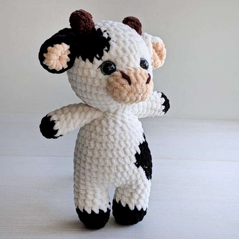 May include: A plush cow toy made of white, black, and brown yarn. The stuffed animal features a white body with black patches, a tan muzzle with dark brown nostrils, small brown horns, and large black eyes. The limbs end in black yarn.