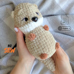May include: A crocheted plush otter toy made of soft beige and white yarn with small brown paws and black plastic eyes. The toy features the text NO SEW in orange, a PDF icon, and a globe icon with the text English, indicating a digital pattern.