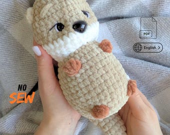 May include: A crocheted plush otter toy made of soft beige and white yarn with small brown paws and black plastic eyes. The toy features the text NO SEW in orange, a PDF icon, and a globe icon with the text English, indicating a digital pattern.