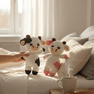 May include: Two handmade crochet cow plush toys. One cow is white with black spots, black ears, and black hooves. The second cow is white with pink spots, pink ears, and pink hooves. Both plushies feature dark eyes and light tan muzzles.