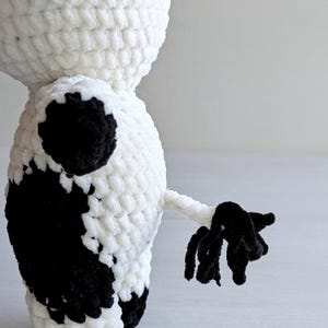 May include: A plush cow toy made of white and black yarn with a crochet texture. The figure features a rounded body, a small tail with a black tufted end, and distinct black patches on the side and back. The yarn has a soft, fuzzy appearance.