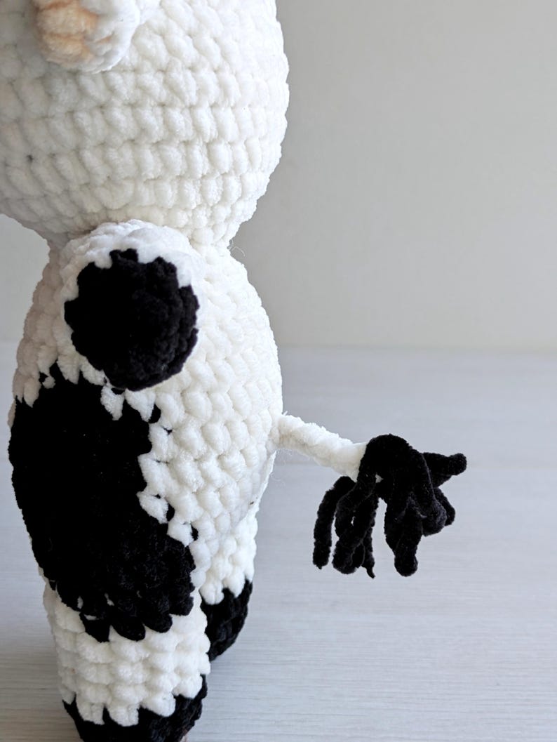 May include: A plush cow toy made of white and black yarn with a crochet texture. The figure features a rounded body, a small tail with a black tufted end, and distinct black patches on the side and back. The yarn has a soft, fuzzy appearance.