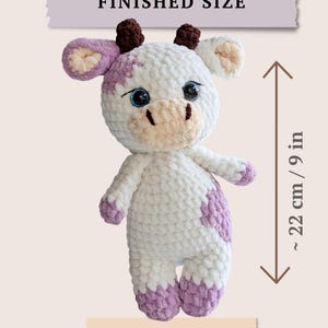 May include: A white and lavender crocheted cow plush toy with dark brown horns, large blue eyes, and a light beige muzzle. The toy features a finished size of approximately 9 inches, as indicated by the text and measurement graphic.