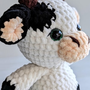 May include: A plush cow toy made of white, black, and brown yarn. The cow features a white body with black patches, a light tan muzzle with dark brown nostrils, dark brown horns, and large, round, glossy black eyes with green accents.
