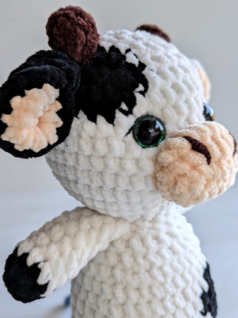 May include: A plush cow toy made of white, black, and brown yarn. The cow features a white body with black patches, a light tan muzzle with dark brown nostrils, dark brown horns, and large, round, glossy black eyes with green accents.