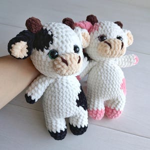 May include: Two handmade crochet cow plush toys made from soft white yarn. One cow features black patches, black ears, and brown horns, while the other features pink patches, pink ears, and brown horns. Both have large black eyes and tan muzzles.