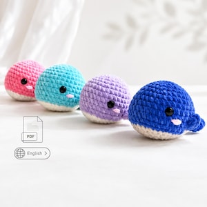 May include: Four round, crocheted whale plushies in pink, turquoise, lavender, and dark blue. Each whale features a white underbelly, black plastic safety eyes, and a small pink stitched mouth. The plushies are arranged in a row on a white surface.