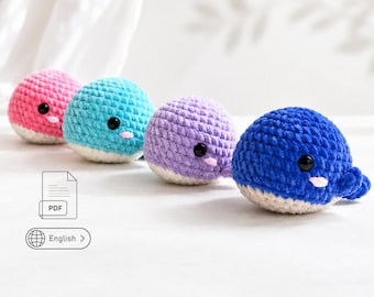 May include: Four round, crocheted whale plushies in pink, turquoise, lavender, and dark blue. Each whale features a white underbelly, black plastic safety eyes, and a small pink stitched mouth. The plushies are arranged in a row on a white surface.
