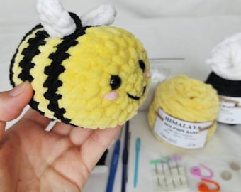 May include: A crocheted bee plush toy with black and yellow stripes, white wings, and pink cheeks. The bee is made with a fuzzy yarn and has a smiling face. The toy is being held in a hand, and there are other crafting supplies in the background.