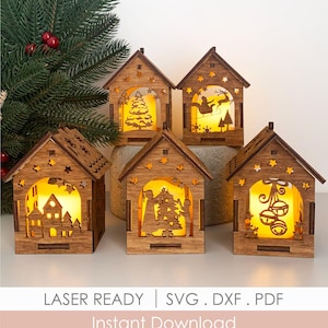 May include: Five laser-cut wooden house-shaped lanterns with warm yellow interior lighting. Each features intricate cutouts of Christmas scenes including a tree, Santa in a sleigh, a village, people decorating a tree, and bells. Text reads: LASER READY | SVG . DXF . PDF Instant Download.