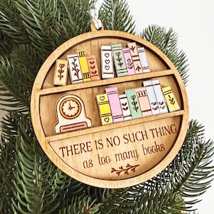 May include: Wooden, round ornament featuring a bookshelf design with colorful books and a clock. The text "THERE IS NO SUCH THING as too many books" is displayed. The ornament is hanging on a Christmas tree.