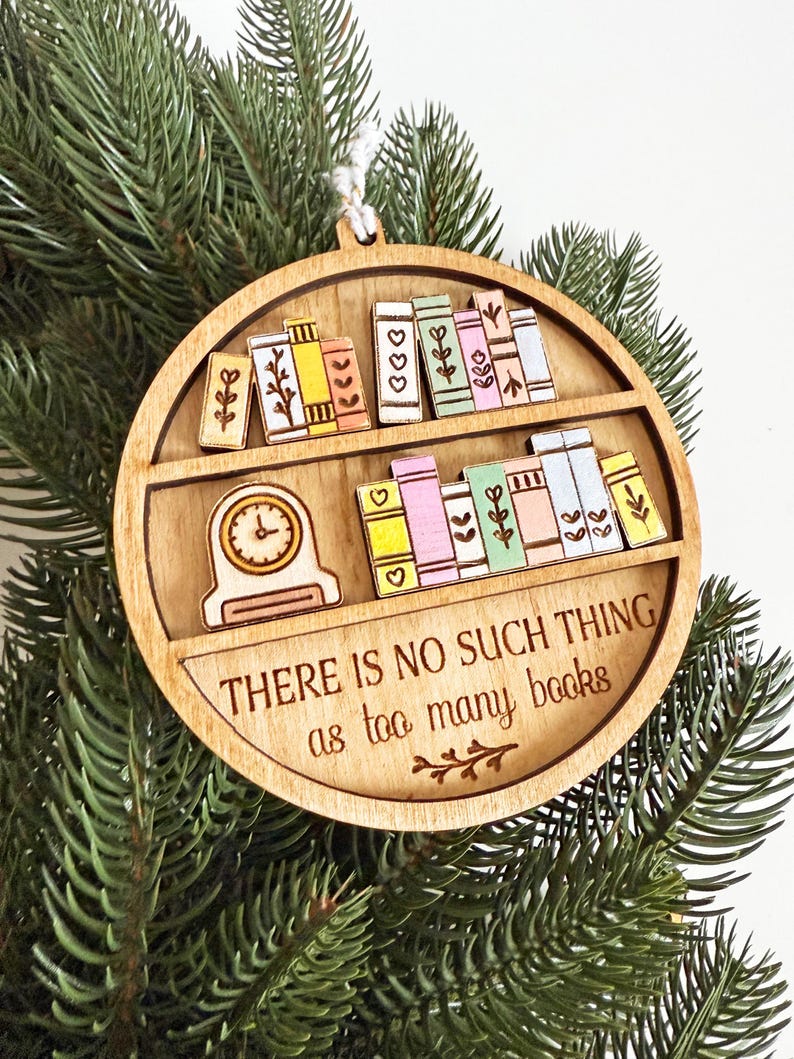 May include: Wooden, round ornament featuring a bookshelf design with colorful books and a clock. The text "THERE IS NO SUCH THING as too many books" is displayed. The ornament is hanging on a Christmas tree.