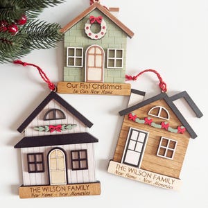 May include: Three wooden house-shaped Christmas ornaments. One is green with a wreath and the text "Our First Christmas in Our New Home." The other two have the text "THE WILSON FAMILY New Home - New Memories."