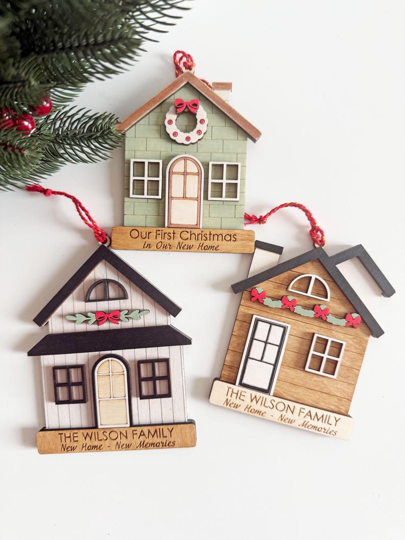 May include: Three wooden house-shaped Christmas ornaments. One is green with a wreath and the text "Our First Christmas in Our New Home." The other two have the text "THE WILSON FAMILY New Home - New Memories."