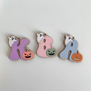 May include: Three wooden letter ornaments shaped as K, B, and A. Each letter is painted in pastel purple, pink, or blue, featuring a small white ghost perched on top and a carved jack-o'-lantern pumpkin in orange or green attached to the side.
