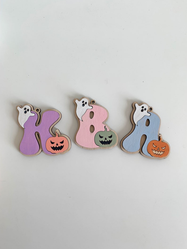 May include: Three wooden letter ornaments shaped as K, B, and A. Each letter is painted in pastel purple, pink, or blue, featuring a small white ghost perched on top and a carved jack-o'-lantern pumpkin in orange or green attached to the side.