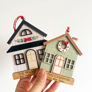 May include: Two wooden house ornaments with red hanging strings. One is white with a black roof and the text "THE WILSON New Home". The other is green with a wreath and the text "First Christmas Our New Home".