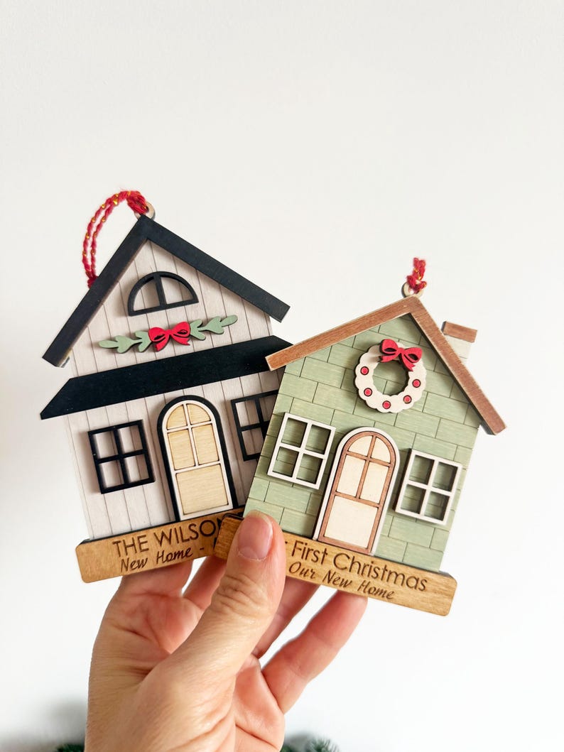 May include: Two wooden house ornaments with red hanging strings. One is white with a black roof and the text "THE WILSON New Home". The other is green with a wreath and the text "First Christmas Our New Home".