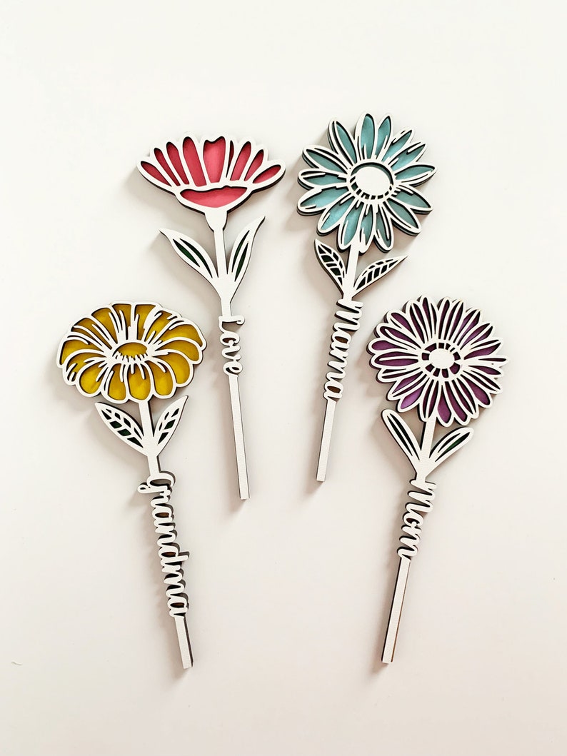 May include: Four wooden flower cutouts with different colored flowers and the words "Love", "Grandma", "Mom", and "Mama" written on the stems.